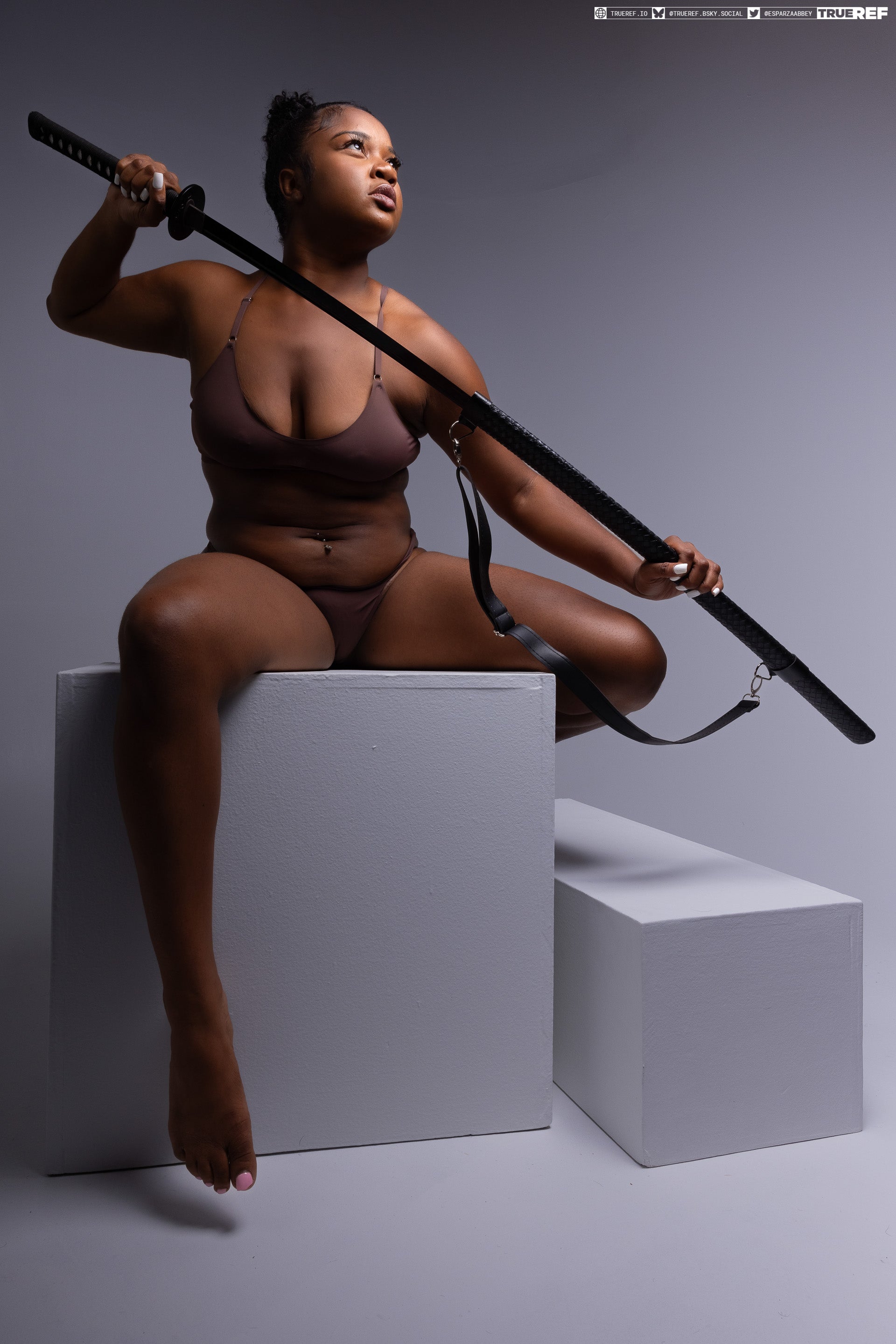 a darkskinned woman sits on a white cube, posing with a katana. she is wearing underwear.