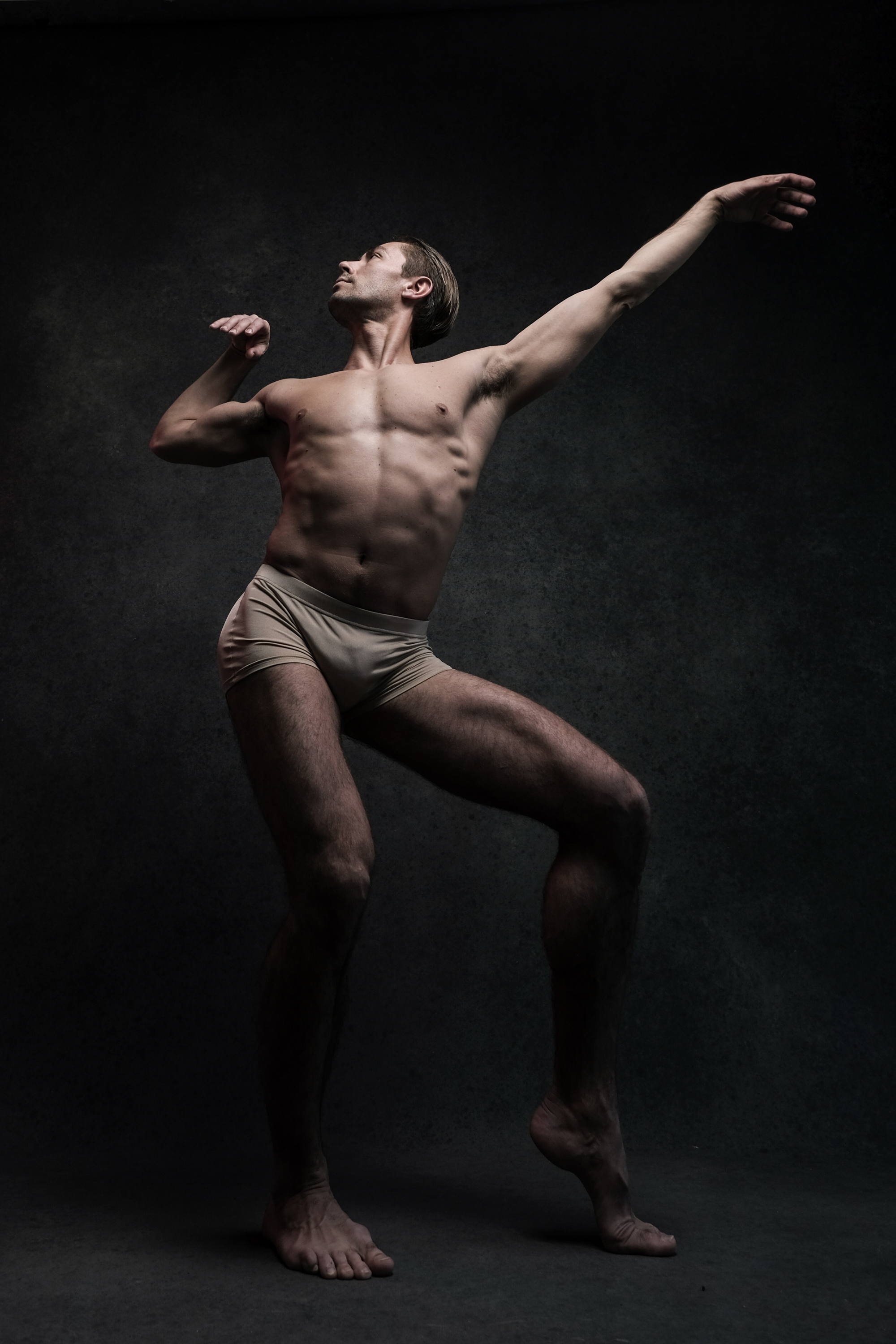 a muscular male dancer wearing beige briefs strikes a dramatic pose against a dark gray background, illuminated by a spotlight. the dancer's body is contorted, demonstrating flexibility and strength. his left arm is extended gracefully overhead while his right arm is bent at the elbow. his right leg is extended outward and his left leg supports his weight. his gaze is directed upwards.