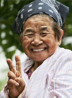 a closeup portrait of an elderly asian woman wearing a bandana, smiling and giving a peace sign with her right hand.
