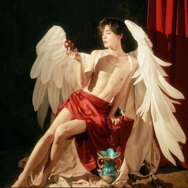 a man with white angel wings is wearing a red skirt. he is posing in a studio with a red background. he is holding an apple in his hand.