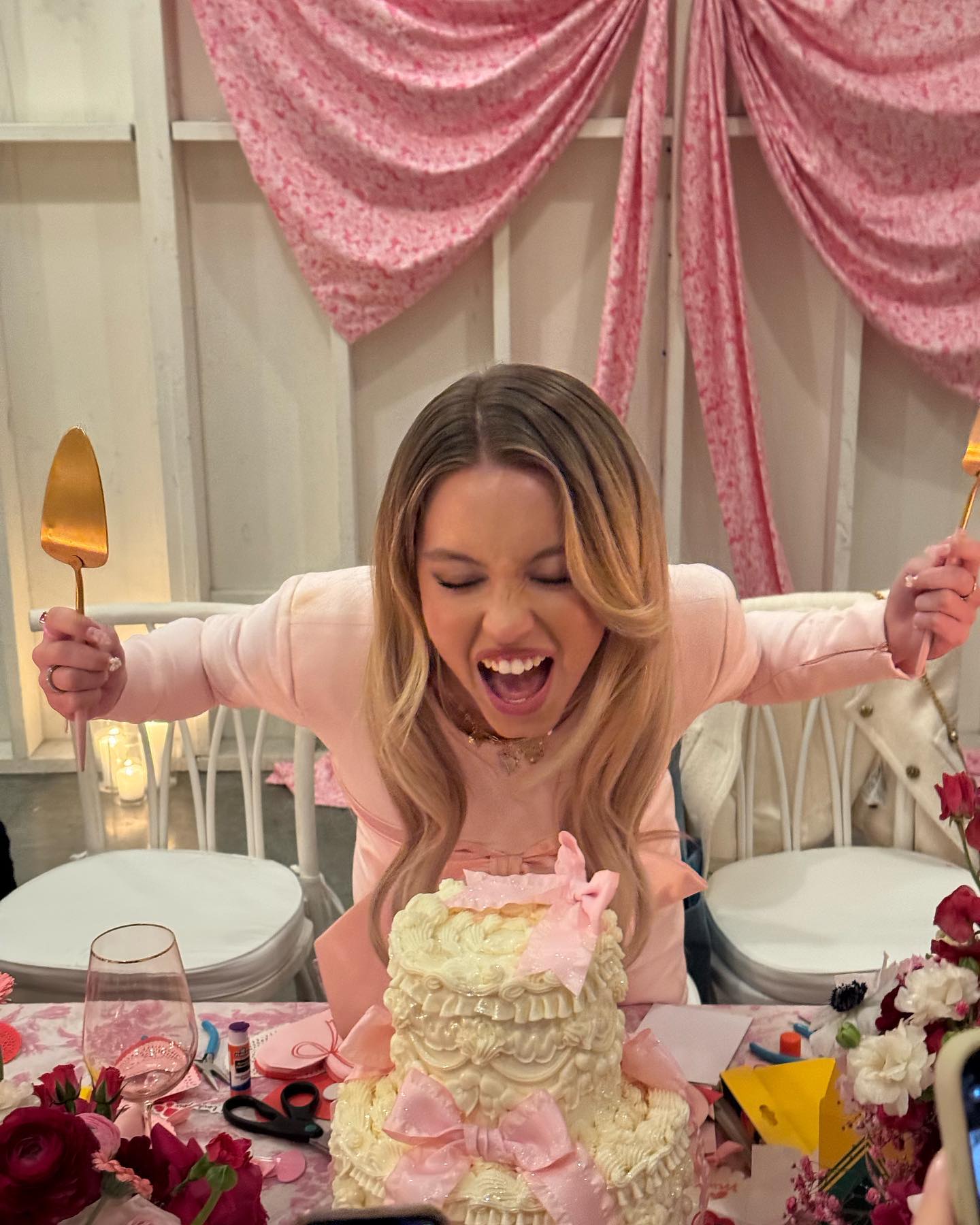 a blonde woman wearing a pink blazer is excitedly cutting into a twotiered, white cake decorated with pink bows and ruffles. the scene is set at a pinkthemed party with pink decorations and flowers visible on the table.  she is holding gold cake servers and her expression is one of pure joy.  the overall feel is of a celebratory, sweet, and fun event.