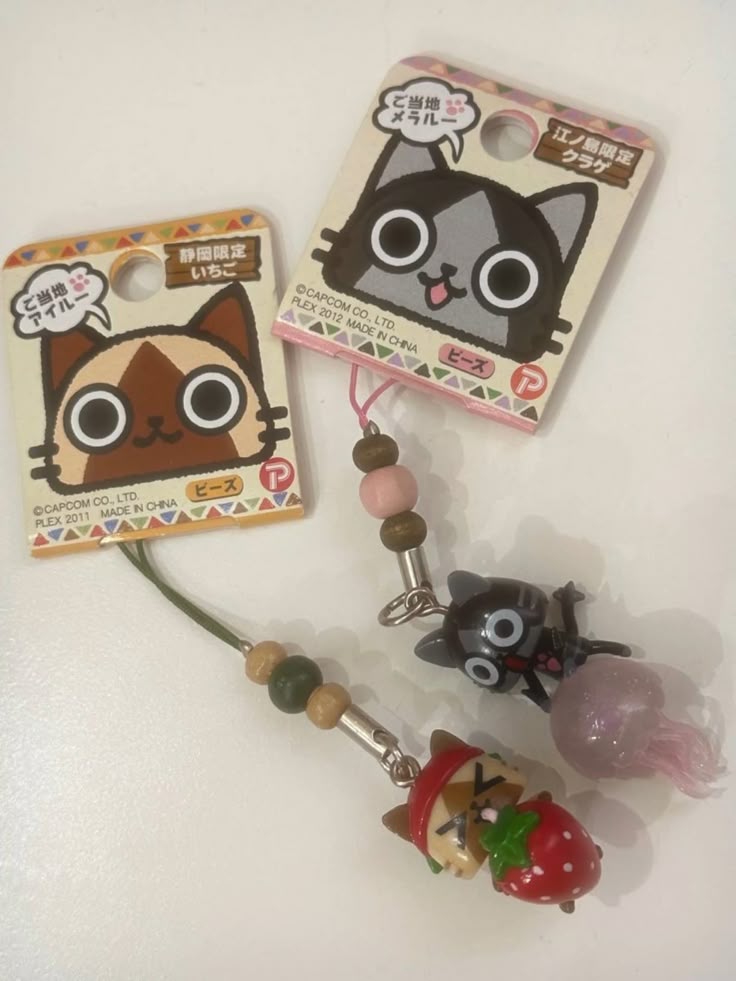 the image shows two monster hunter themed keychains. one keychain features a brown and white ailu holding a strawberry, which indicates it's a shizuokalimited "local souvenir" keychain. the other keychain features a black and gray merarou with a jellyfish, indicating it's a enoshimalimited "local souvenir" keychain.