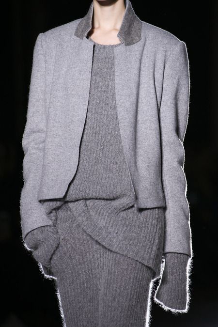 a model wearing a grey sweater and a grey jacket is walking down a runway. she has a serious expression on her face and is wearing a simple, yet elegant outfit.