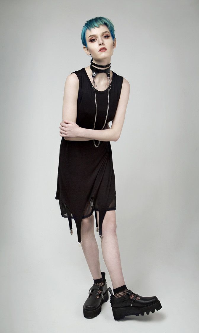 a full shot of a woman posing in a black dress and chunky black platform shoes. she has short blue hair and is wearing a black choker necklace. the background is a solid gray. the image is clean and welllit. the woman's pose is slightly angled, and she is looking directly at the camera. the style is goth or alternative.