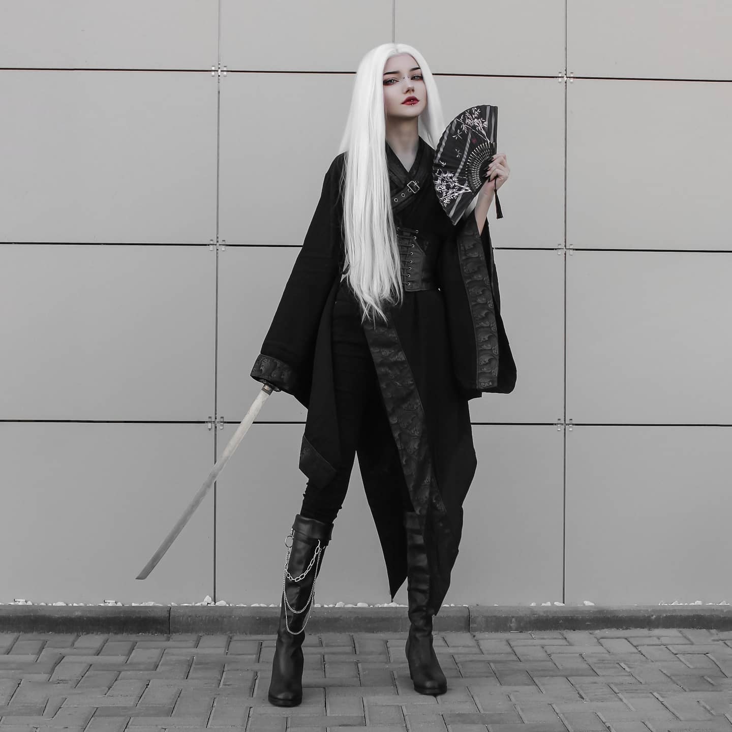 a full shot of a woman in a black and white cosplay outfit.  she's wearing a long black kimonostyle coat over black pants, high black boots with chains, and has long, straight white hair. she holds a black hand fan and a katana (samurai sword) and stands against a grey wall. the overall aesthetic is dark, edgy, and elegant. the image is in monochrome, enhancing the dramatic effect.