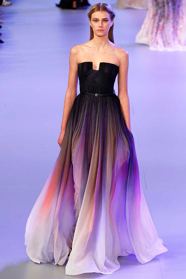 a fashion model walks the runway in a black, ombre gown that transitions from black to purple to white. she has long straight hair and is wearing a simple black belt. the dress is flowing and elegant.  the runway is white and the background is blurred, making the model the focus.