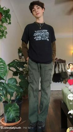 a young man is standing indoors, he is wearing a black longsleeve shirt with white text on it. he is also wearing green cargo pants and black shoes. he has a necklace on with a black cord. he is looking at the camera.