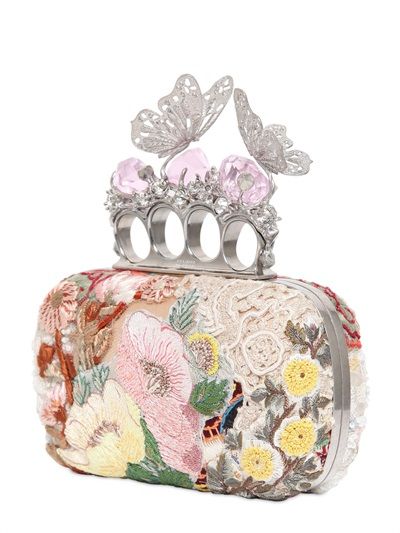 a closeup shot of a clutch purse with a decorative silver handle featuring butterfly accents and pink gemstones. the purse itself is ornately embroidered with floral patterns. the overall impression is of a highfashion, elegant accessory.