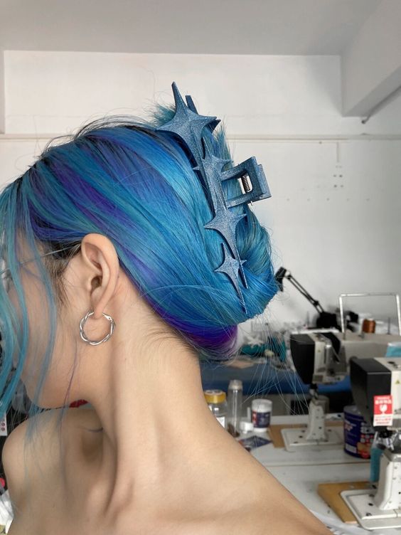 a woman with bright blue and purple hair with a large blue hair clip, showing the side of her face, wearing silver hoop earrings. she is looking away from the camera and is standing in front of a row of sewing machines.