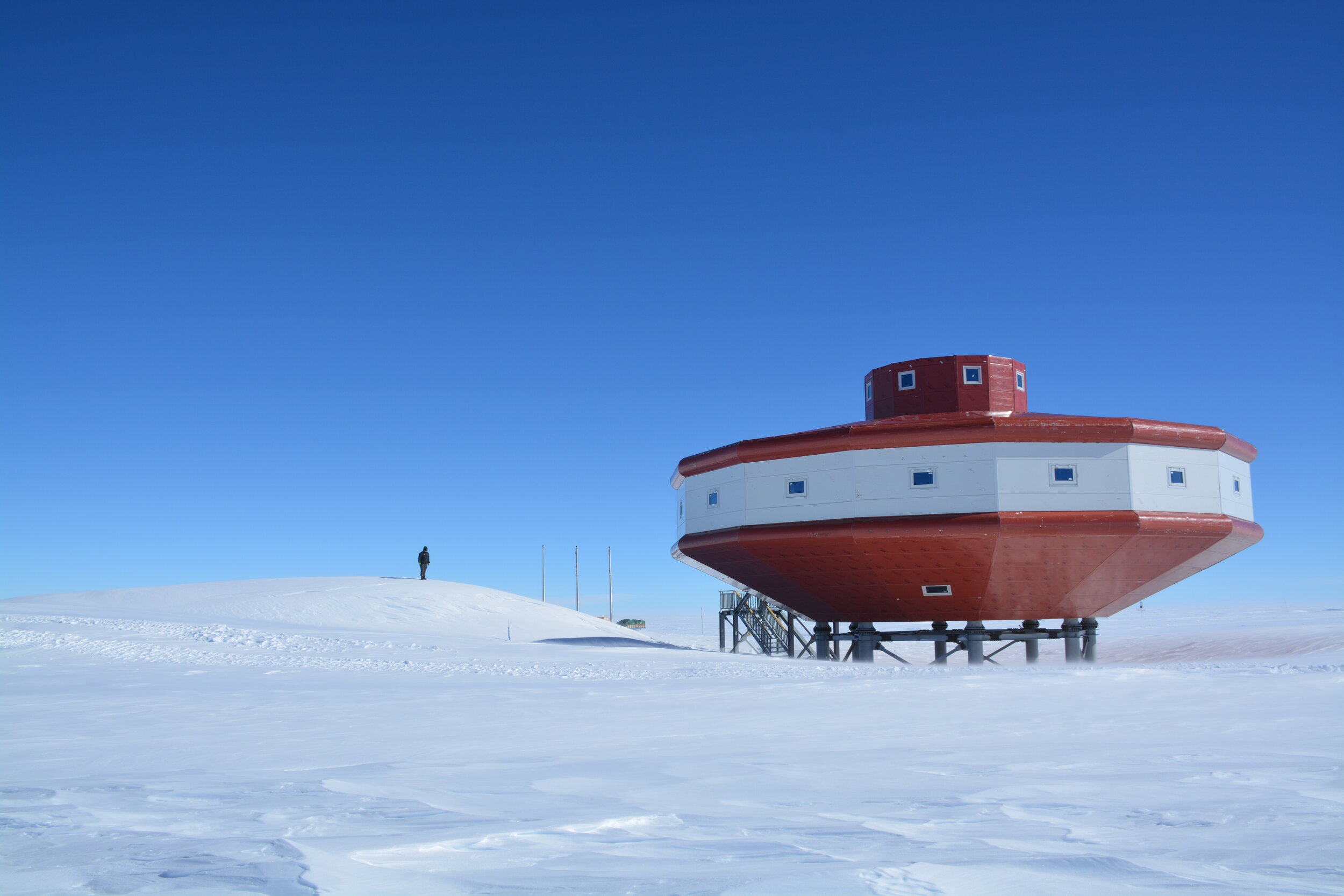 a red and white building with a unique, circular shape is situated on a snowy landscape under a clear blue sky. the building appears to be a research station in antarctica. the image is taken from a distance, showing the building's isolation and the vastness of the snowy terrain. the horizon is visible in the background, and the entire scene is characterized by the harsh beauty of the antarctic environment.