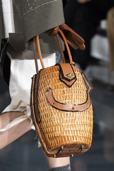 a person wearing a gray jacket and white shorts is holding a woven basket handbag with brown leather details. the person is wearing a brown glove.