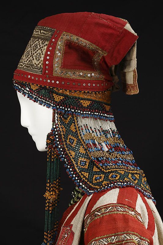 a detailed studio shot features a mannequin wearing an elaborate, traditional headdress and garment. the headdress is predominantly red, adorned with various types of beadwork, sequins, and embroidered patterns. the beadwork cascades down the sides of the mannequin's face. the garment beneath is red and white striped with embroidered accents. the headdress's intricate designs and patterns are highlighted by the studio lighting.