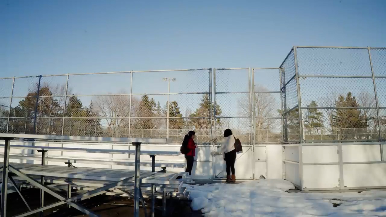 two women are standing on a snowcovered sports field, engaged in conversation.  they are bundled up in winter coats and boots. the field is surrounded by a chainlink fence and there are bleachers in the foreground.  the trees in the background are bare, indicating it is winter. the sun is shining brightly, casting long shadows on the snow.  the overall tone of the image is bright and cheerful.
