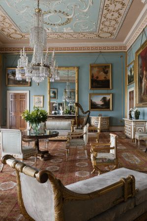 a wide shot of a brightly lit formal living room decorated in a traditional style with pastel blue walls, goldaccented furniture, antique paintings, a large patterned rug, and various decorative objects including a harp, plant, and crystal chandelier.