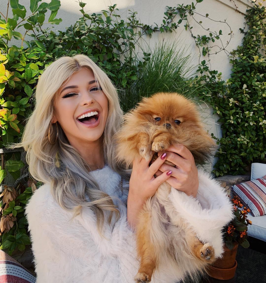 a smiling blonde woman holds a small, fluffy pomeranian dog in her arms outdoors. she is wearing a white sweater and large hoop earrings, and her nails are painted. the background features lush greenery, a white wall, and a glimpse of patio furniture.