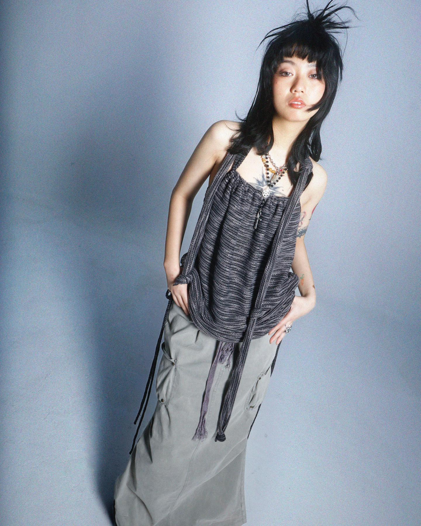 a fulllength studio shot of a young woman with short, dark hair styled with choppy bangs and a slightly spiky texture. she's wearing a long, light grey cargo skirt and a dark grey knit sleeveless top with long ties that hang down.  the top has a relaxed, slightly slouchy fit. she accessorizes with multiple necklaces.  the overall style is edgy and alternative, with a slightly grunge feel. the background is a simple, light grey. the model appears to be of asian descent. the image is welllit and professionally taken, likely for a fashion magazine or editorial.