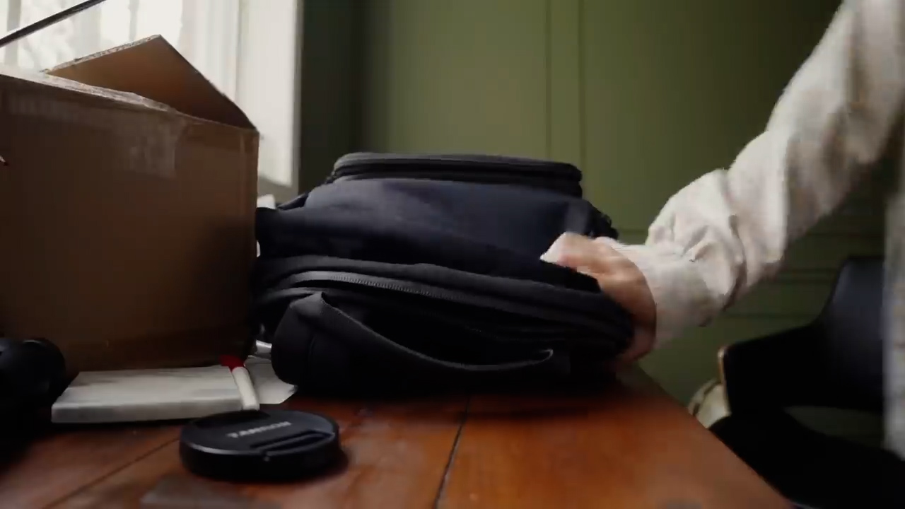 a woman is seen placing a black backpack on a wooden table. there's a cardboard box, paperwork, and a camera lens on the table. the scene is likely indoors with natural light coming from a window to the left.