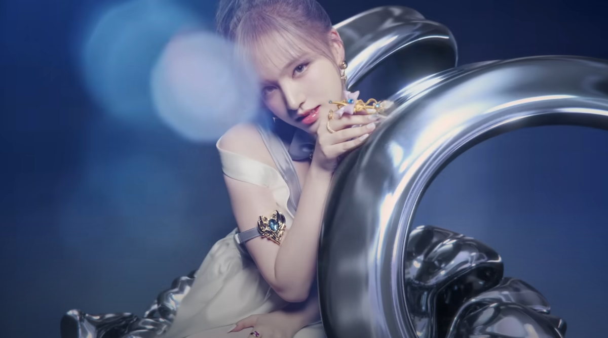 a young woman with blonde hair, likely a kpop idol, is the focus of the image. she's posing against a backdrop of abstract, metallic silver shapes, possibly conveying a futuristic or glamorous concept. her outfit, a flowing white dress, contrasts with the harder lines of the background. she holds a delicate, ornate object in her hand. the lighting is soft and ethereal, contributing to the overall dreamy atmosphere of the image.