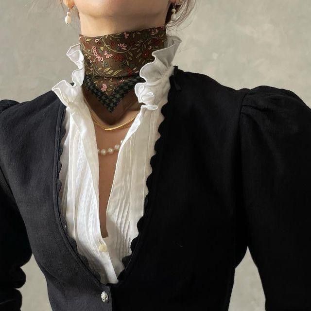 a woman is wearing a black cardigan over a white blouse with a ruffled collar and a floral scarf tied around her neck. she is also wearing a gold necklace, a pearl necklace, and pearl earrings. the style appears to be vintage or dark academia. the image shows the woman's upper body.