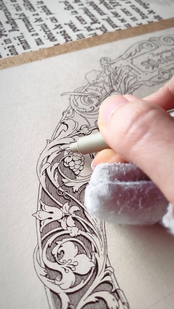 a closeup shot shows a person's hand carefully drawing intricate details onto a piece of paper. the drawing depicts an ornate design with floral and scrollwork elements, reminiscent of vintage or antique illustrations. the person is using a lightcolored pen, and their hand is gently resting on a piece of fabric. the drawing is on a pale background, possibly paper or parchment,  and rests on a page of an antique book with visible text. the image showcases a precise and meticulous artistic process.
