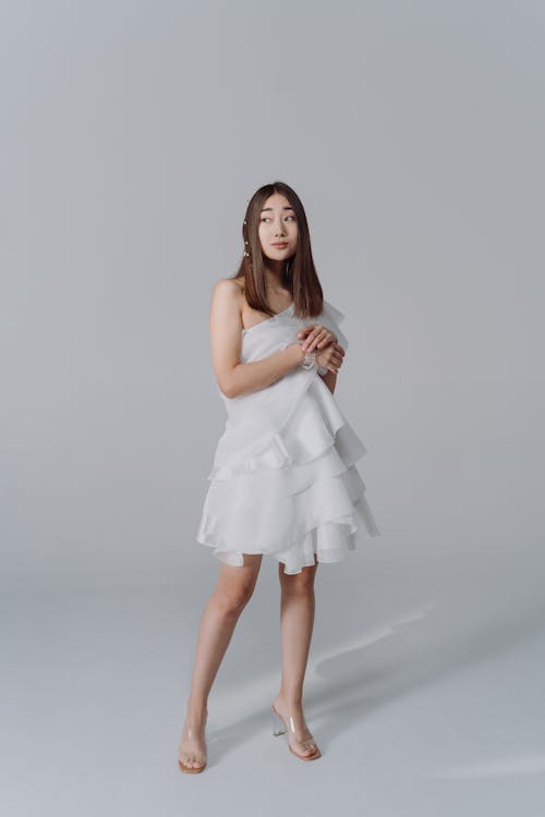 the image shows a young asian woman posing in a white, ruffled oneshoulder mini dress.  she's wearing clear highheeled sandals and has her hair styled in a long, straight style. her pose is relaxed yet elegant, and her expression is somewhat pensive. the background is a plain, light gray studio backdrop. the overall aesthetic is minimalist and clean.