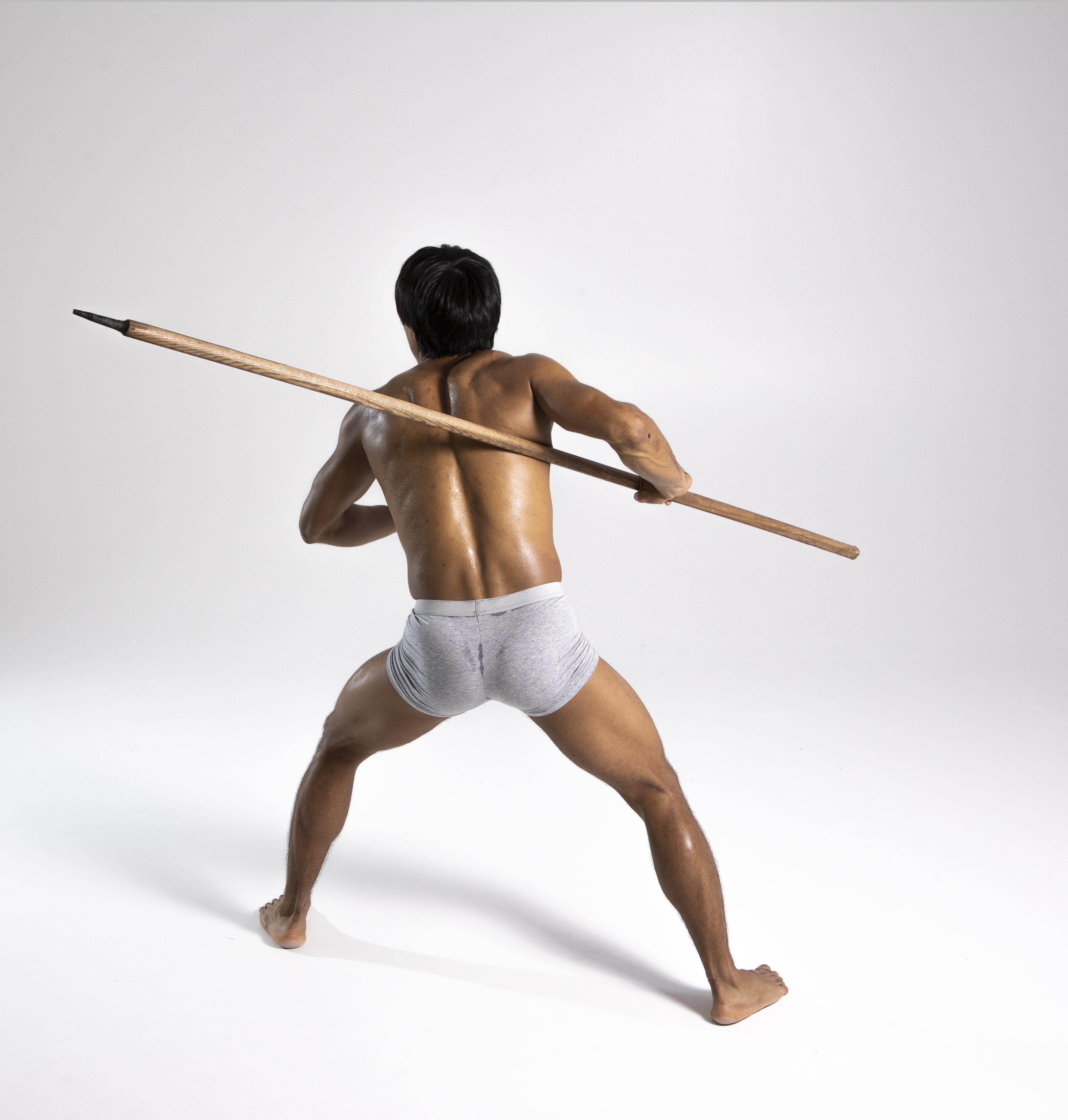 a shirtless man with an athletic build is standing barefoot on a white background, holding a spear. he is viewed from the back, wearing a loincloth. he is in a warrior pose, gripping the spear with both hands, one behind him and the other in front. the spear is a long, wooden shaft with a dark pointed tip. the lighting is bright, casting a shadow behind the man.
