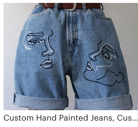 this is an image of custom handpainted denim shorts featuring a line art drawing of two faces. the shorts are blue denim with a brown leather belt and are laid flat against a white background. the art on the shorts is black and white.