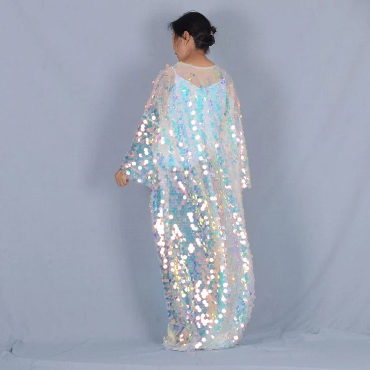 a woman is modeling a long white iridescent sequin maxi dress with long sleeves.  she is standing in front of a light blue background.