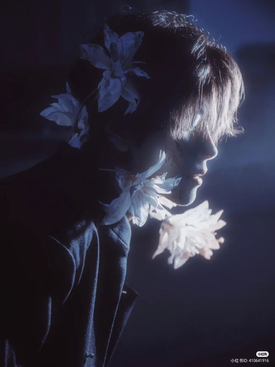 a portrait of a man with flowers in his hair. the image has a dark and moody atmosphere with blue lighting. the man is looking away from the camera and his face is partially obscured by shadows.