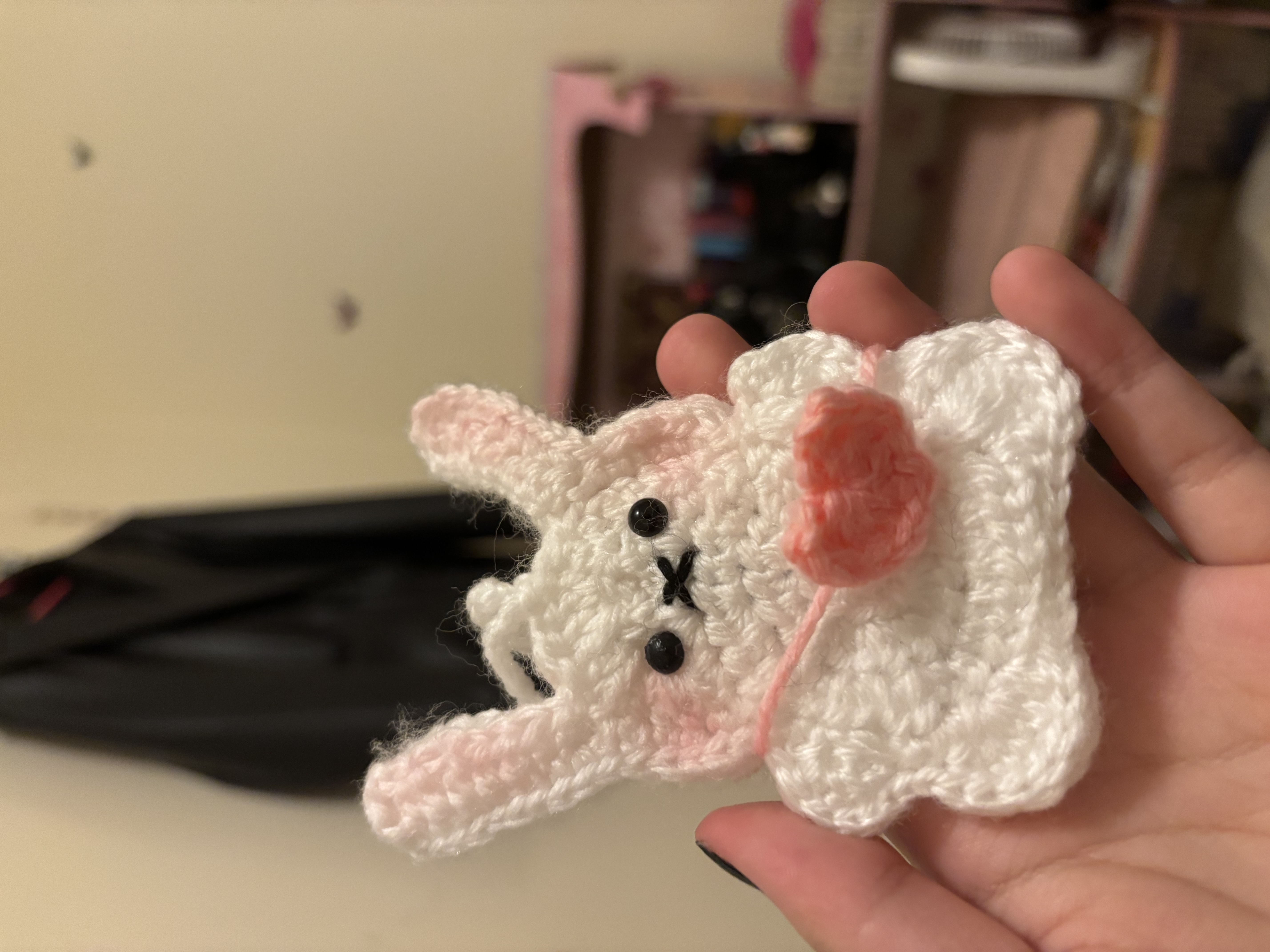 a closeup shot of a handmade crocheted white bunny with pink accents held in someone's hand. the bunny has pinklined ears, pink blush on its cheeks, and a small pink heart hanging from a string around its neck. the background is blurry, showing a dark jacket hanging on a wall and a pink shelf or storage unit. the focus is on the handcrafted bunny.