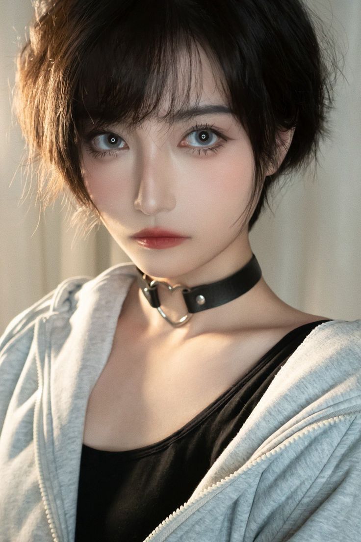 a closeup portrait of a young asian woman with short black hair wearing a grey hoodie and a black tank top. she is looking directly at the camera with a slight smile on her face. she is wearing a black choker with a silver heart pendant. the background is a white wall.
