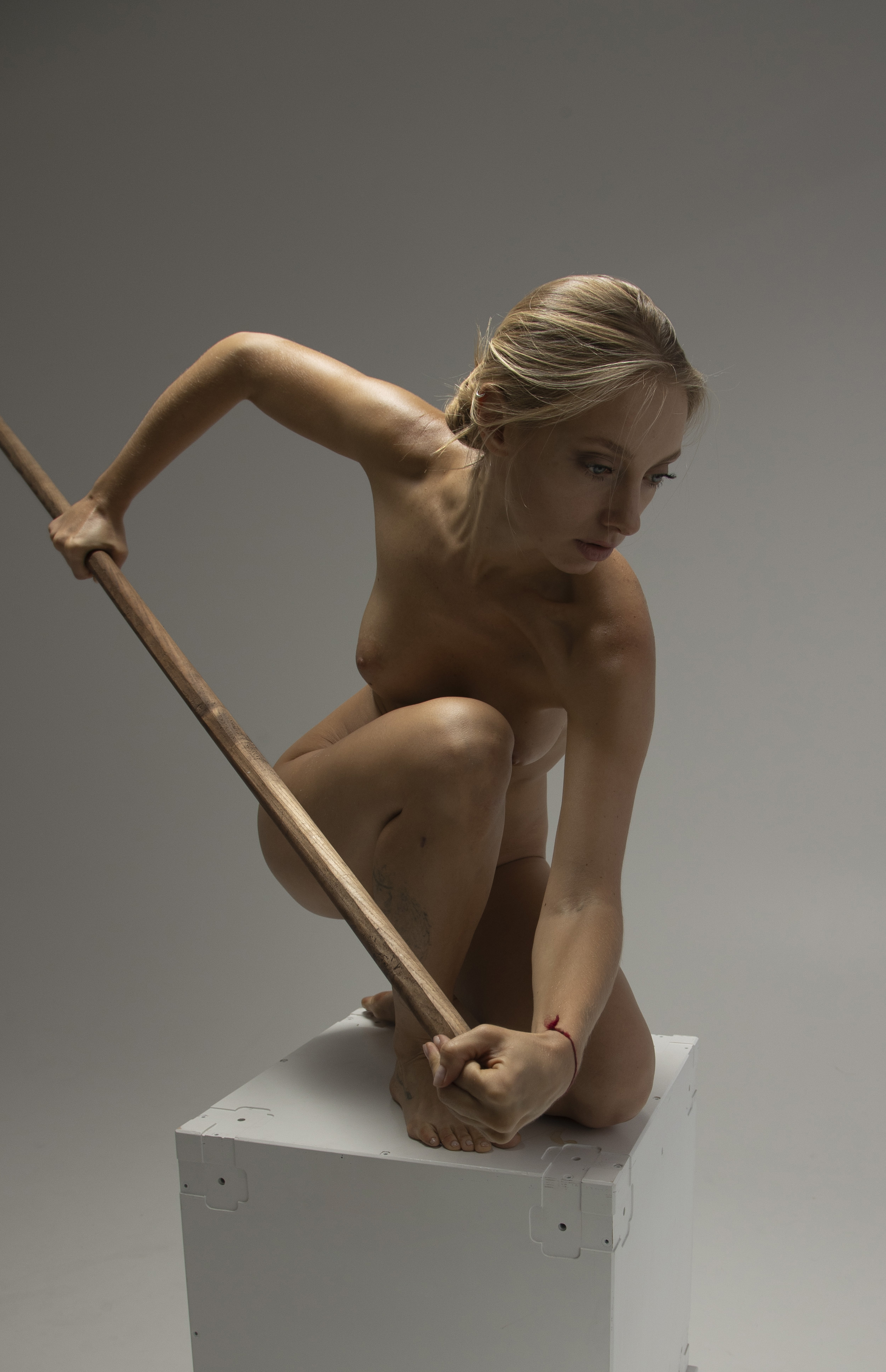 a nude blonde woman is squatting on a white cube while holding a wooden pole. she has bare feet and is wearing a bracelet on her right wrist. there is a tattoo on her left thigh. the background is a plain grey studio backdrop.
