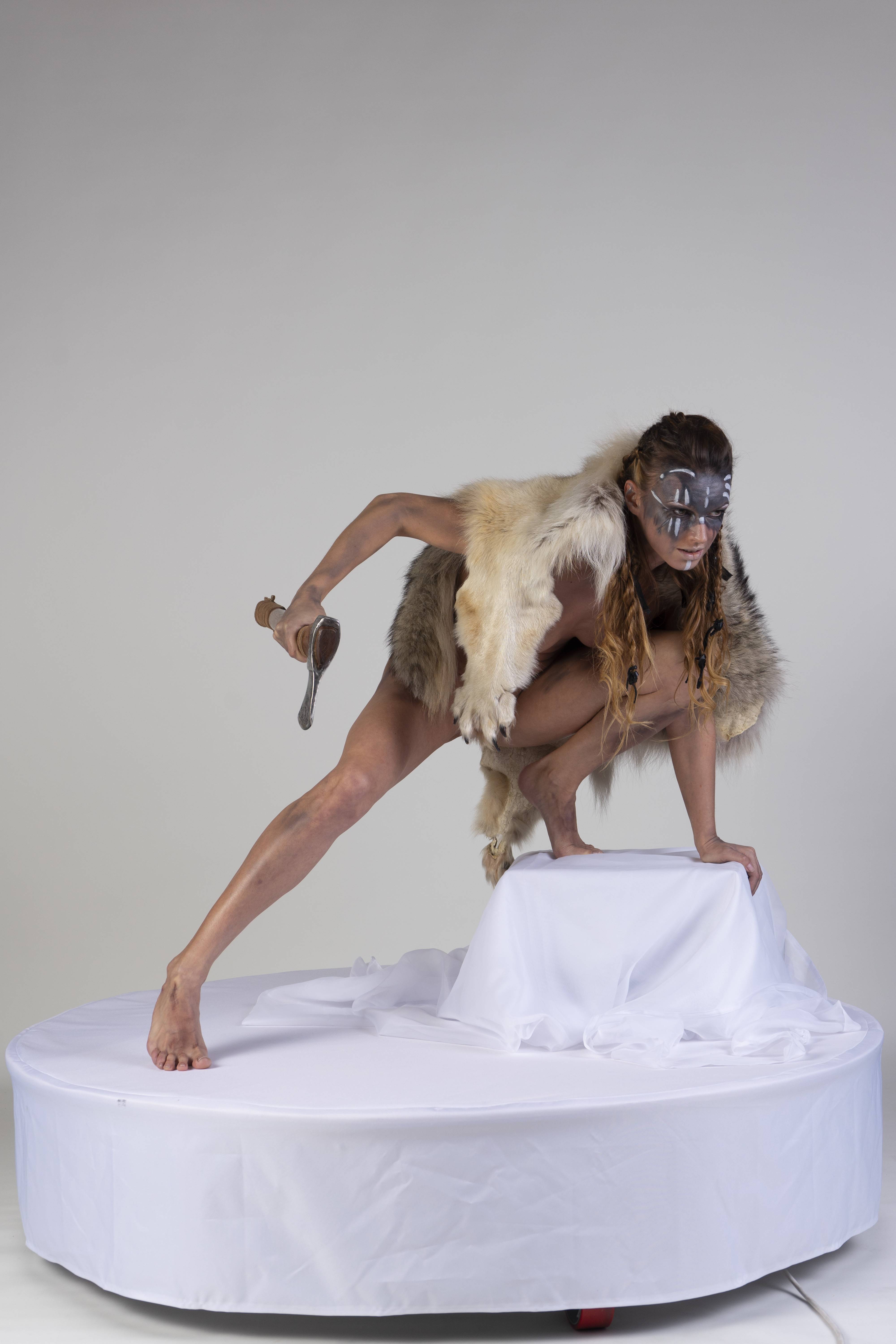a woman dressed as a warrior in a fur costume and face paint is squatting on a white platform. she is holding an axe in her right hand.