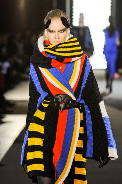 a fashion model walks the runway in a brightly colored outfit. her outfit is predominantly black, yellow, red, and blue.  she is wearing a long, striped coat with a large, colorful scarf. the model is wearing a black belt with a large, black buckle. she is also wearing black hair pieces.