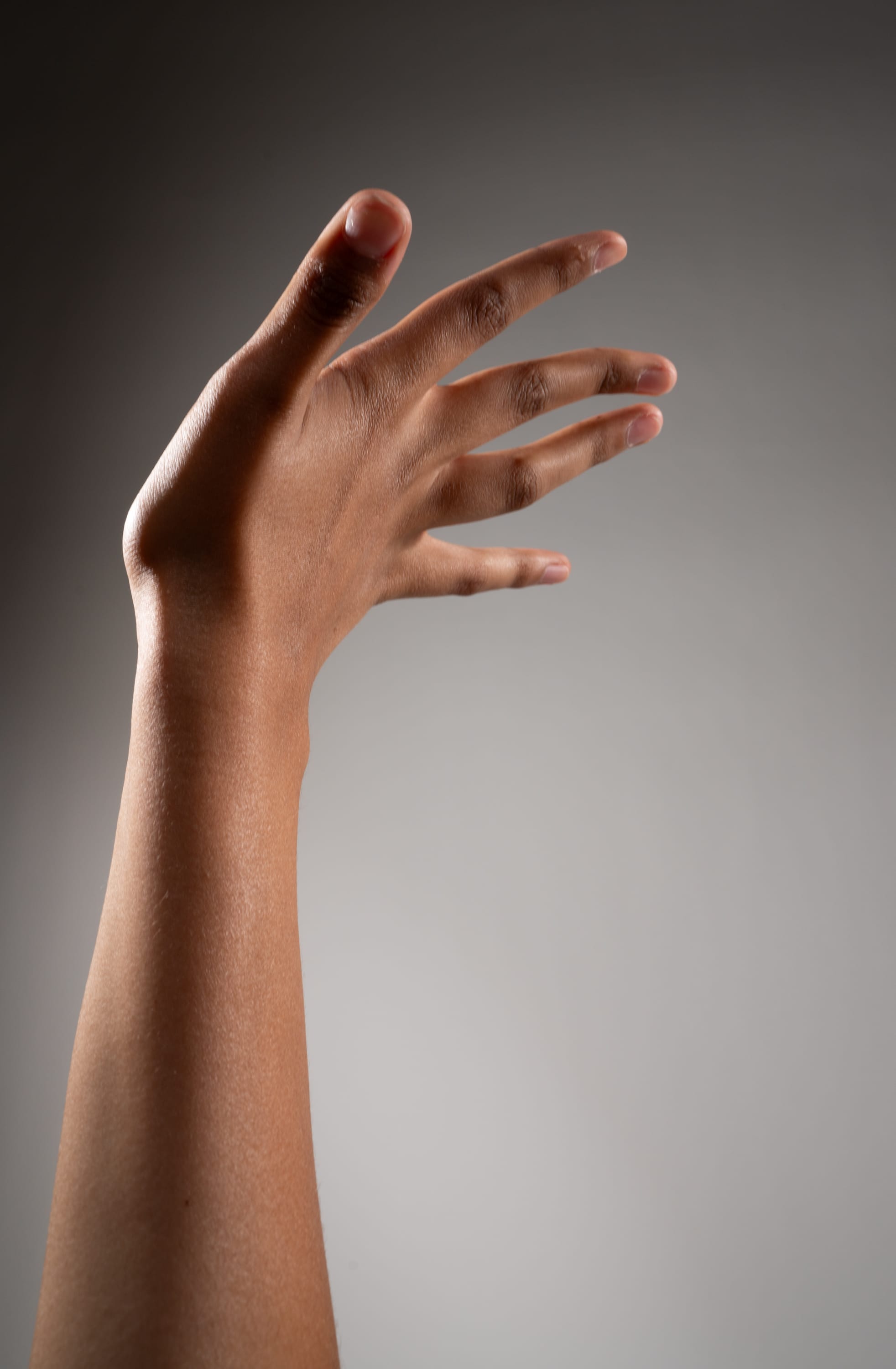 this image is of a person's left hand and arm extended upward against a gray background. the hand is open with the fingers spread apart. the lighting is soft and diffused, creating a smooth and even tone on the skin. the image is taken from a side view, emphasizing the length and curve of the arm.