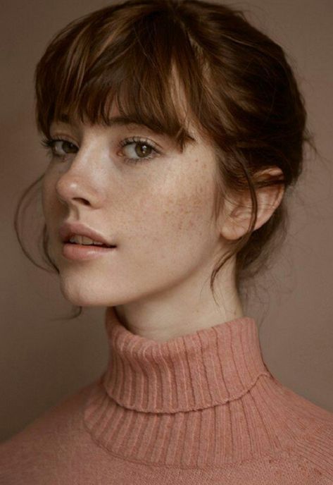 the image is a closeup portrait of a young woman with light skin and freckles. she has shoulderlength brown hair with straight bangs that partially cover her forehead.  her hair is styled in a slightly messy bun. she is wearing a ribbed, dusty rose or peachcolored turtleneck sweater. the lighting is soft and even, casting a subtle glow on her face. the background is a muted, neutral beige. the overall mood is calm, serene, and elegant. the woman's expression is gentle and slightly pensive, conveying a sense of quiet introspection.