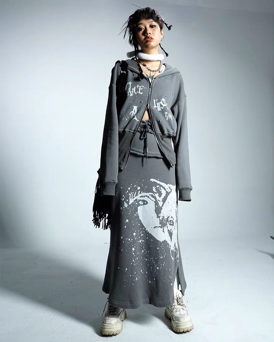 a fulllength studio shot of a young woman modeling a grey oversized hoodie and a matching long skirt with a graphic print. she is wearing white sneakers and simple accessories. the overall style is casual streetwear with a trendy and contemporary feel. the background is plain and minimalistic, allowing the clothing to take center stage.