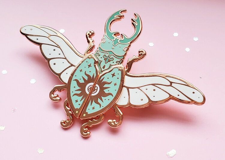 a closeup studio shot of a decorative beetle enamel pin. the pin is mostly turquoise, white, and gold, and it has wings, legs, horns, and a split shell with a sun design. the background is pink and scattered with white sequins.