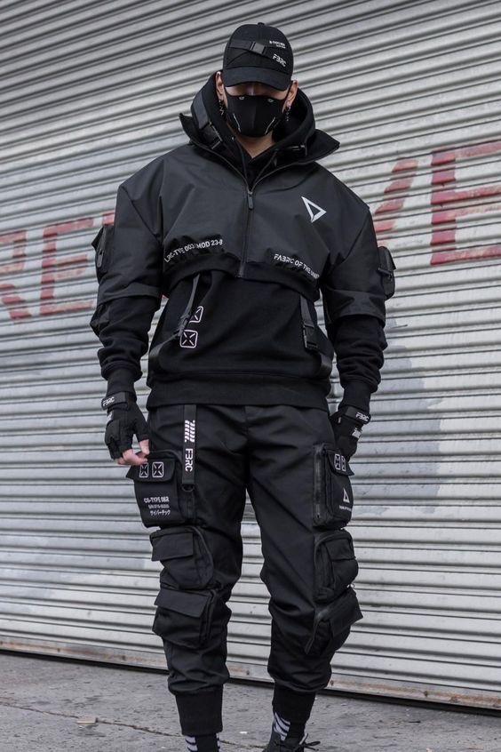 a man in a black outfit stands in front of a metal garage door. he is wearing a black jacket with white details, black cargo pants with white details, a black hoodie, a black mask and a black cap.