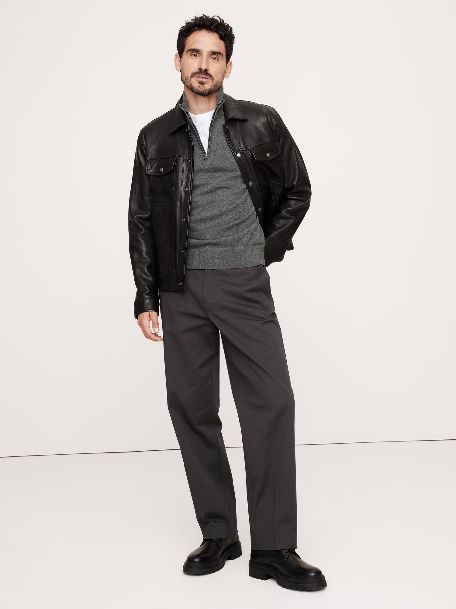 a man stands in a studio wearing a black leather shirt jacket over a gray quarterzip sweater, gray wide leg pants, and black boots. he is facing forward with his hands at his sides. the background is plain white.