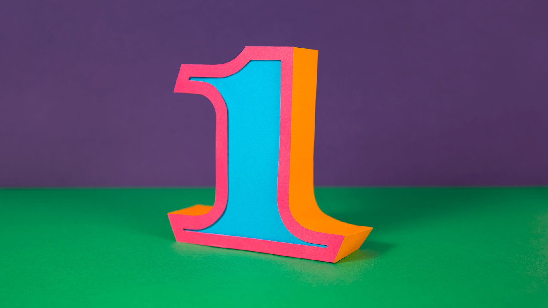 the image shows the number "1" crafted out of paper and standing on a green surface. the number is threedimensional and brightly colored with a pink outline, an orange face, and a blue inset. the background is a twotone purple and green, adding to the vibrant and playful aesthetic.