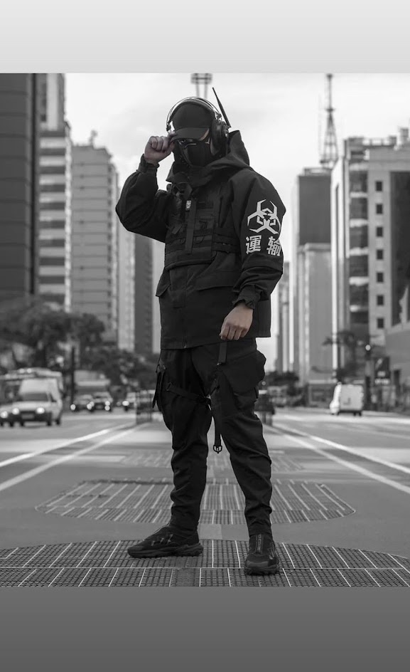 a man dressed in black, including a jacket, pants, and boots, stands on a city street. he is wearing a mask and has a serious expression. the background features tall buildings and a sense of urban grit. the man's attire and pose convey a cool, futuristic, and perhaps even slightly dystopian vibe.