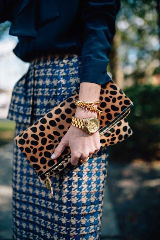 closeup of a woman wearing a stylish outfit. she is wearing a navy blue blouse and a blue and white houndstooth skirt. she's carrying a leopard print clutch and wearing gold accessories, including a watch and bracelets.  the image focuses on the details of her clothing and accessories, highlighting the textures and patterns. the background is blurred, drawing attention to the woman and her attire.