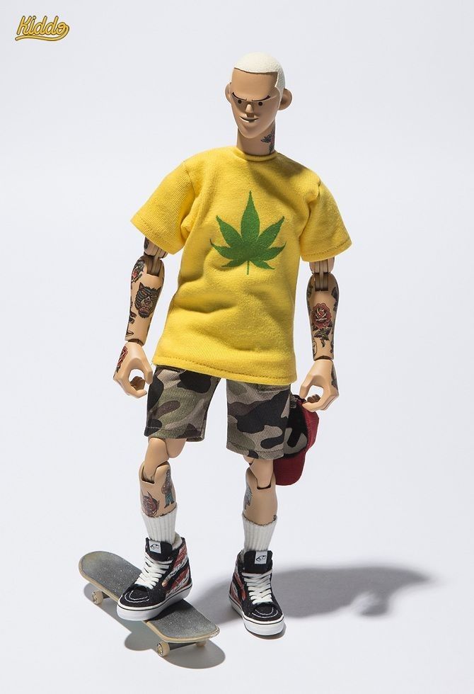 a plastic action figure of a male skater wearing a yellow tshirt with a green marijuana leaf on it, camo shorts, red and black vans high tops and a red cap. the figure has tattoos on his arms and is holding a skateboard in his hand. it is standing on a white background.