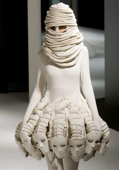 a model is on a runway wearing a white dress with a sculptural design.  the dress is made of many layers of fabric that are folded and shaped into masks and other abstract forms. the model's face is covered with a white mask that is made of fabric.  the dress is very avantgarde and futuristic, creating a surreal and whimsical aesthetic.  the model's white dress is a powerful statement of individuality and style.