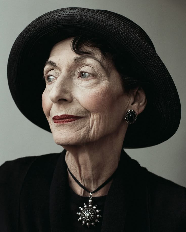 a portrait of an elderly woman wearing a black hat and black clothing. she has wrinkles and red lipstick, and is wearing a necklace and earrings. she is looking to the side. the image evokes elegance and sophistication.
