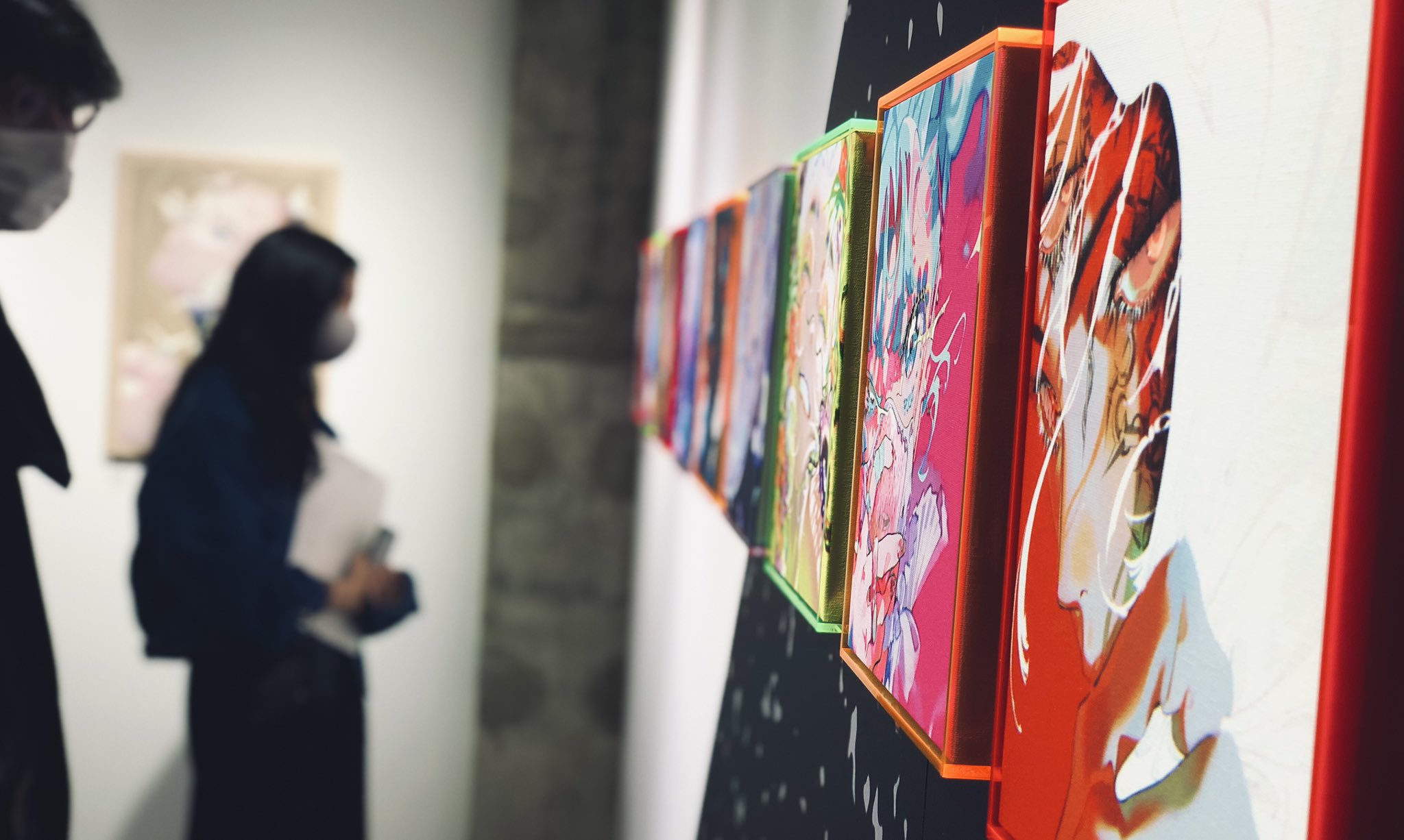 the image shows a closeup view of a gallery wall featuring a series of colorful, abstract artworks framed in red and black. the art pieces are predominantly pink, blue, and yellow, with bold lines and abstract forms. two people, their faces obscured by masks, are out of focus in the background, suggesting they are observing the art. the gallery is welllit with artificial lighting, creating a bright and modern atmosphere.