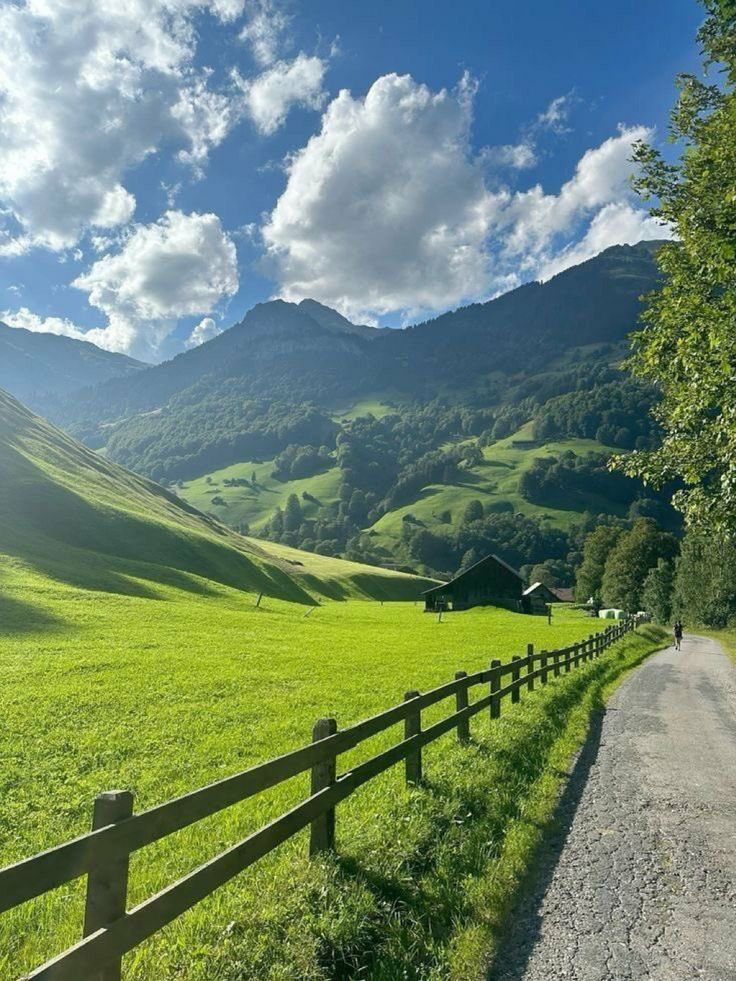 a scenic landscape featuring a winding path beside a wooden fence, with a lush green field extending towards rolling hills and mountains under a blue sky with fluffy clouds. a barn is nestled in the field, and trees line the edges of the path.