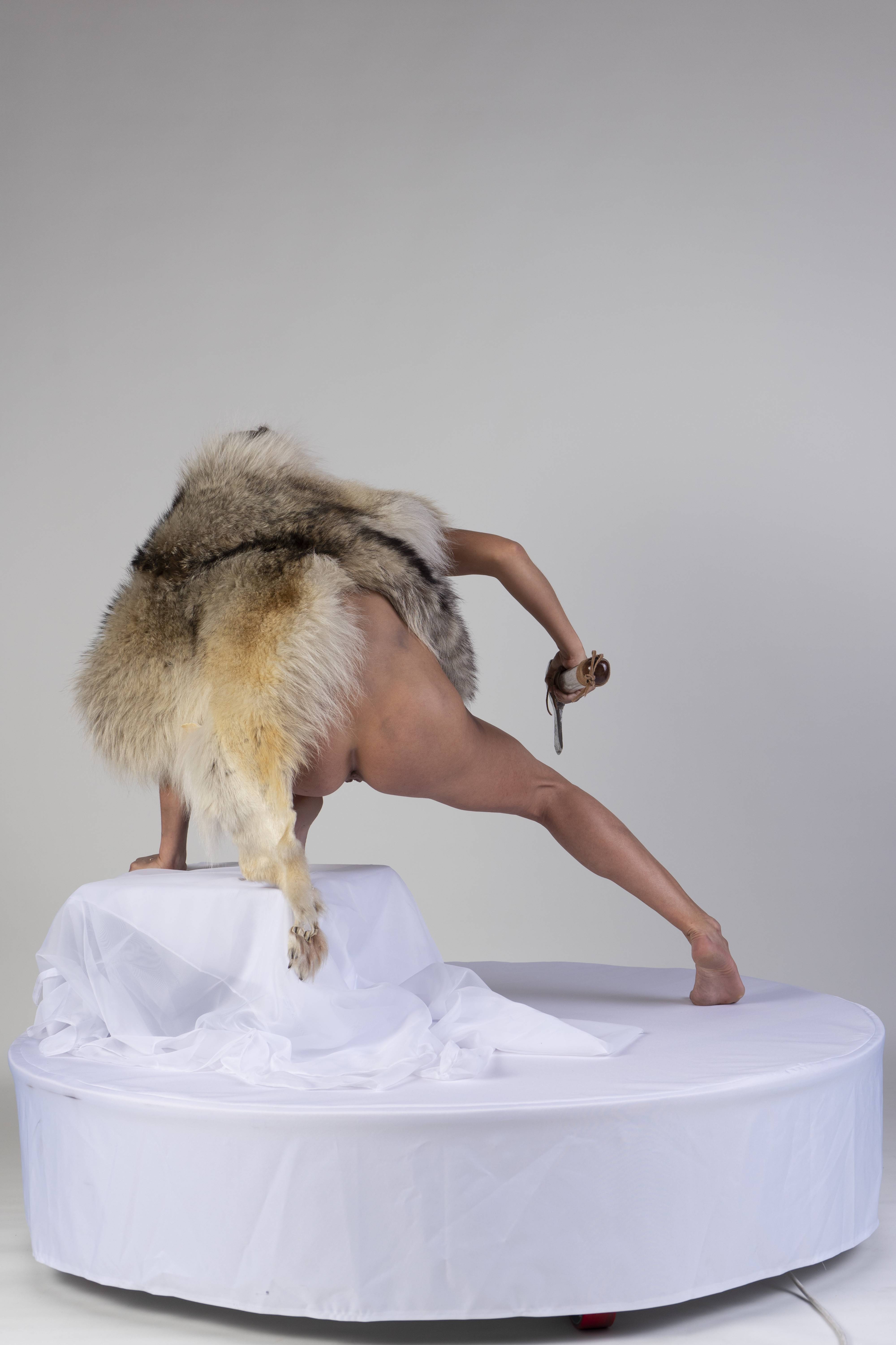 a nude woman wearing a fur pelt is standing on a white platform, doing yoga.
