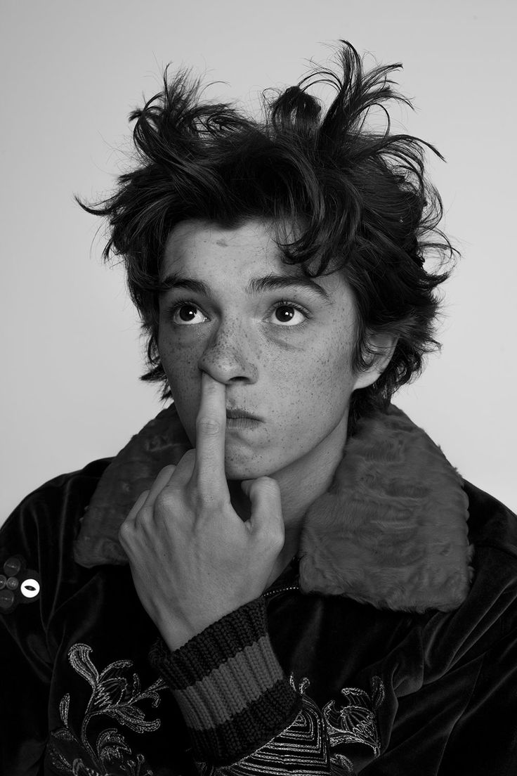 a black and white portrait of a young man wearing a black velvet jacket with a fur collar. he has a thoughtful expression as he touches his nose with his index finger. he has short dark hair, and his eyes are looking upwards.